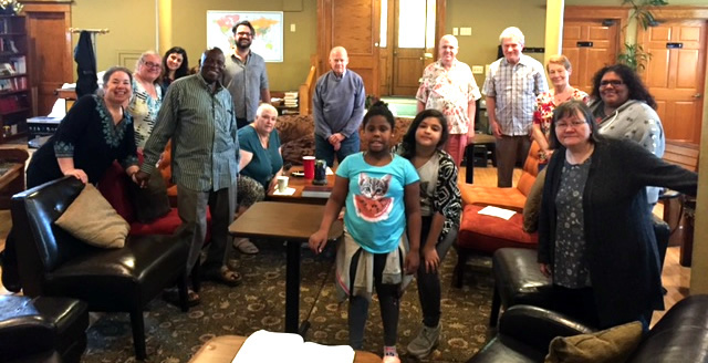 Group photo of diverse community members of all ages gathered in a warm community space with yellow walls.